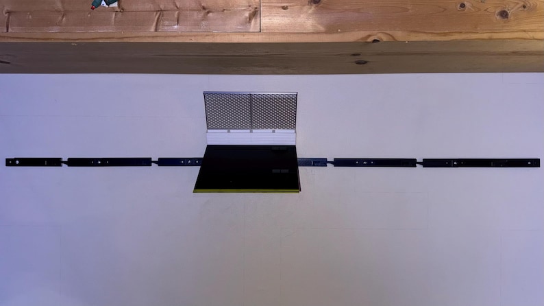 May include: A black and silver range hood with a rectangular design. The hood has a mesh screen and a black panel. It is mounted on a white wall with black support bars extending to the sides.