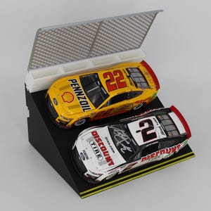 May include: Two miniature race cars, one yellow with the number 22 and the other white with the number 2, are displayed on a black angled stand. The yellow car has "PENNZOIL" branding. A white and gray barrier is behind the cars.
