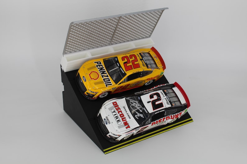 May include: Two miniature race cars, one yellow with the number 22 and the other white with the number 2, are displayed on a black angled stand. The yellow car has "PENNZOIL" branding. A white and gray barrier is behind the cars.