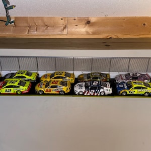 May include: A collection of colorful toy race cars displayed on a shelf. The cars feature various sponsor logos and racing numbers. The shelf has a metal fence design, and the cars are arranged in a row, showcasing their vibrant paint schemes.