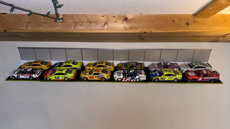May include: A collection of colorful toy race cars displayed on a shelf. The cars feature various sponsor logos and racing numbers. The shelf has a metal fence design, and the cars are arranged in a row, showcasing their vibrant paint schemes.