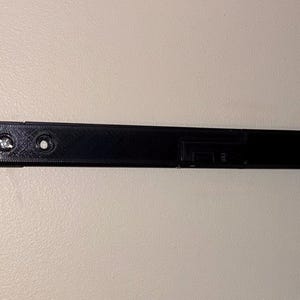 May include: A black, rectangular object with screw holes and screws. The object appears to be made of plastic or a similar material. The object is mounted on a light-colored wall. The object is likely a component of a larger item.
