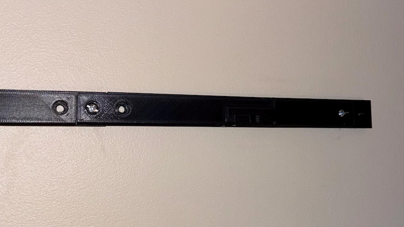 May include: A black, rectangular object with screw holes and screws. The object appears to be made of plastic or a similar material. The object is mounted on a light-colored wall. The object is likely a component of a larger item.