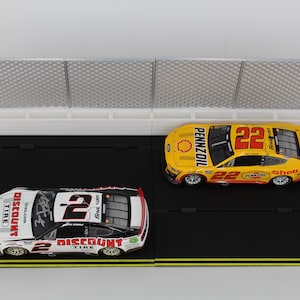 May include: Two toy race cars, one white and red with the number 2, and one yellow with the number 22, sit on a black track with a white fence. The cars have sponsor logos.