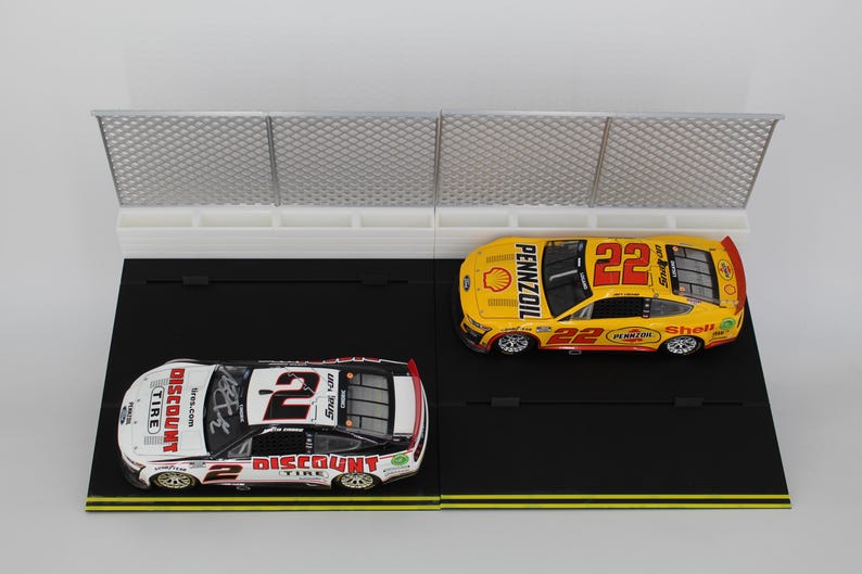 May include: Two toy race cars, one white and red with the number 2, and one yellow with the number 22, sit on a black track with a white fence. The cars have sponsor logos.