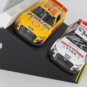 May include: Two model race cars, one yellow and one white, are displayed on a black angled stand. The yellow car has the number 22 and Pennzoil branding. The white car features Discount Tire branding and a signature.
