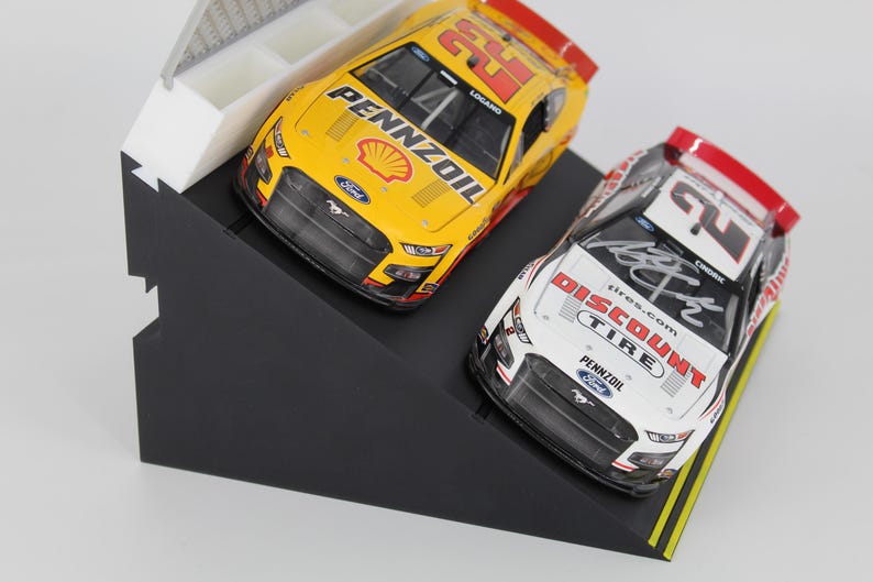 May include: Two model race cars, one yellow and one white, are displayed on a black angled stand. The yellow car has the number 22 and Pennzoil branding. The white car features Discount Tire branding and a signature.