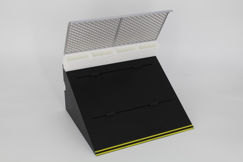May include: Black display stand with a slanted design, featuring a white top section with four compartments and a gray, grid-like lid. The base is black with yellow stripes along the bottom edge. Ideal for showcasing jewelry or small items.