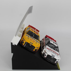 May include: Two toy race cars, one yellow and one white, are displayed on a black angled stand. The yellow car has "Pennzoil" branding. A white structure with steps is behind the cars. The cars are angled as if racing.