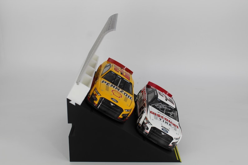May include: Two toy race cars, one yellow and one white, are displayed on a black angled stand. The yellow car has "Pennzoil" branding. A white structure with steps is behind the cars. The cars are angled as if racing.