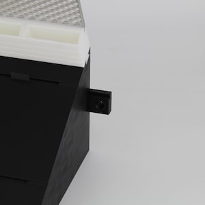 May include: A black and white storage container with a textured top and a small black latch. The container has multiple compartments and a layered design. The background is plain white.
