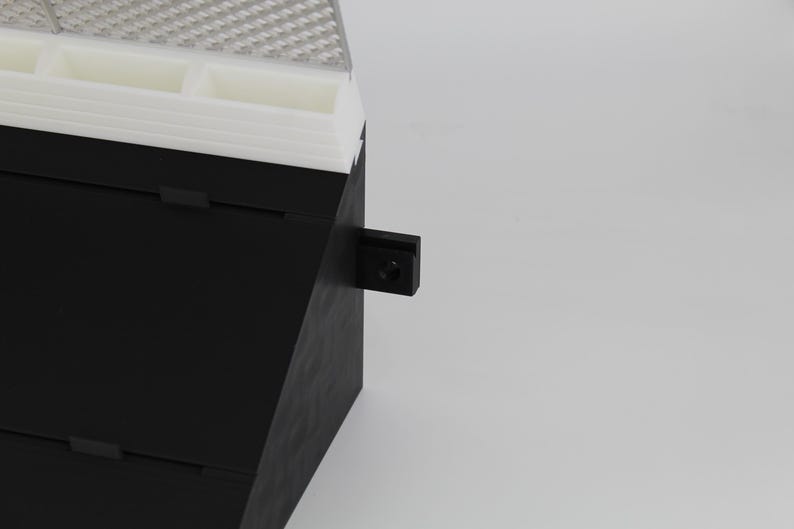 May include: A black and white storage container with a textured top and a small black latch. The container has multiple compartments and a layered design. The background is plain white.