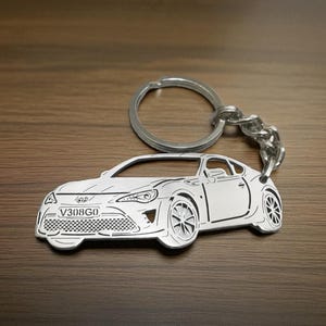 May include: Silver-colored car-shaped keychain with a key ring and chain. The keychain features a detailed outline of a sports car, including the wheels, headlights, and license plate with the text "V308G0".
