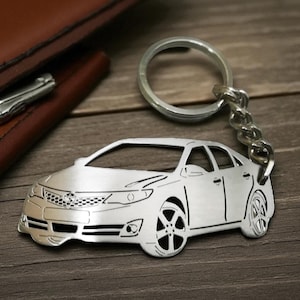 May include: Silver car-shaped keychain with a key ring and chain. The keychain is next to a brown leather wallet on a wooden surface. The car design includes details of the headlights, grill, and wheels.