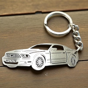 May include: Silver car-shaped keychain with a key ring and chain. The keychain features a detailed outline of a car with the license plate reading "M1LRLTE". The car has detailed wheels and a front grill.