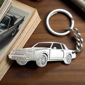 May include: Silver car-shaped keyring with a chain and ring. The keyring has the text "GBODYBOLO" on the front. The keyring is on a wooden surface next to a vintage car photo.