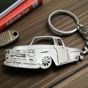 May include: Silver-colored metal keychain in the shape of a classic Chevrolet pickup truck. The keychain has a key ring and chain. The truck has the name "Edgar" on the license plate.