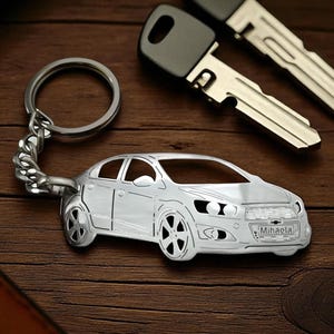 May include: A silver car-shaped keychain with a nameplate that reads "Mihaela" is displayed next to two car keys. The keychain is attached to a metal ring and chain. The items are on a wooden surface.