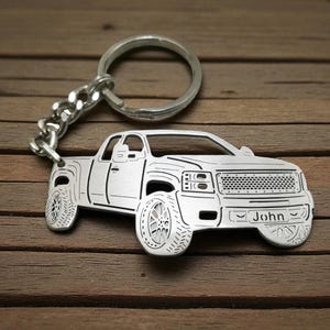 May include: Silver-colored truck keychain with a large key ring and chain. The truck design includes detailed tires and the name "John" engraved on the front. The keychain is displayed on a wooden surface.