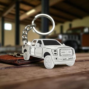 Puede incluir: Un llavero de metal blanco con un contorno detallado de una camioneta. El llavero está unido a una cadena y un anillo plateados. La camioneta tiene el texto "iam_jwh1" en la parte delantera. El llavero descansa sobre una cartera de cuero marrón.