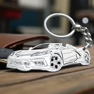 May include: A silver car-shaped keyring with a chain. The keyring features a detailed outline of a sports car with the text "JC1PARA" on the front. The keyring is resting on a wooden surface.