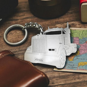 May include: Silver-colored semi-truck keychain with the name "Alexis" engraved on the front. The keychain is attached to a silver ring and chain. The truck is detailed with headlights, a grill, and wheels. The keychain rests on a wooden surface with a wallet and map.