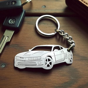May include: A silver car-shaped keychain with a key and car keys on a wooden surface. The keychain features a detailed outline of a car. The car keys are black. The background is a dark brown wooden surface.