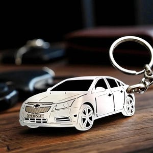 May include: A silver-colored car-shaped keychain with a key ring. The keychain is a detailed outline of a car, with the word "PHIN-E" visible on the front. The keychain is resting on a wooden surface.