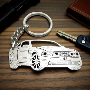 May include: Silver car-shaped keychain with a chain and ring. The keychain features a detailed outline of a car with the number "44" on the front. Other items, including keys and a wallet, are in the background.