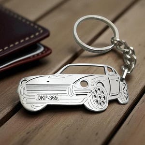 May include: Silver car-shaped keychain with a key ring and chain. The keychain features a detailed car design with visible wheels and a license plate that reads "DKP-369". The keychain is on a wooden surface next to a brown leather wallet.