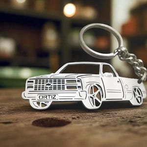 May include: A white metal keychain in the shape of a pickup truck. The keychain has a detailed design, including the word "ORTIZ" on the front. The keychain is attached to a silver chain and ring.