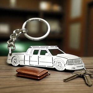 May include: A silver-colored car-shaped keychain with a key ring and chain. The keychain is next to a brown leather wallet and a set of keys on a wooden surface. The car design features a truck with detailed cutouts.