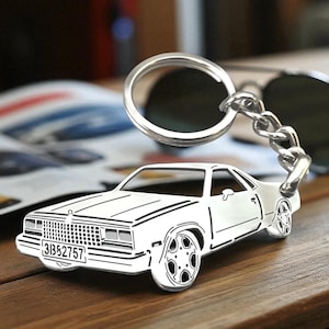 May include: A silver car-shaped keychain with a license plate that reads "31352757". The keychain is attached to a chain and a keyring. Sunglasses are in the background on a wooden surface.