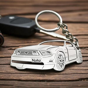 May include: Silver car-shaped keychain with the name "Nabz" engraved on the front. The keychain is attached to a key ring and chain, with a car key and a black leather key holder in the background. The car has detailed wheels and a front grill.