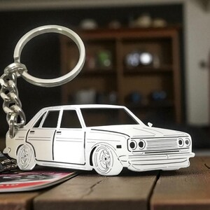 May include: A silver-colored car keychain with a detailed outline of a classic sedan. The keychain includes a large ring and a chain. The car design features headlights, a grille, and wheels. The keychain rests on a wooden surface.