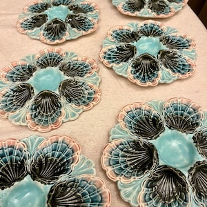 May include: Six ceramic oyster plates with a scalloped shell design. Each plate features a central well and six surrounding compartments. The plates are in shades of blue, pink, and black, with a glossy finish. A decorative tableware item.