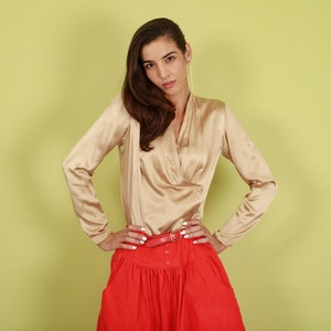 May include: A woman wearing a beige satin blouse with a red skirt and a brown belt. She is standing in front of a light green background.