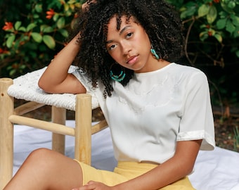 May include: A woman with curly dark hair sits on a white blanket with a wooden stool beside her. She is wearing a white embroidered top with a yellow skirt and turquoise earrings.