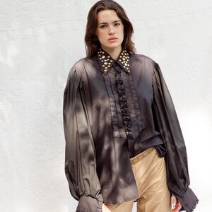 May include: A dark brown long-sleeved shirt featuring a ruffled front placket, wide cuffs, and a pointed collar embellished with gold-toned studs and clear crystals. The shirt is paired with light tan trousers that have a metallic gold waistband.