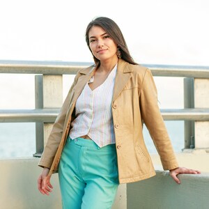 May include: A woman wearing a tan leather jacket over a white and pastel striped shirt and teal blue pants. She is standing in front of a metal railing with a body of water in the background.
