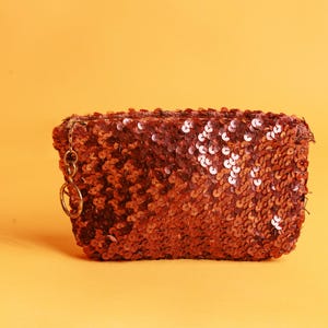 May include: A rectangular, sequined clutch in a warm, reddish-brown hue. The bag is covered in small, reflective sequins, creating a shimmering effect. A small gold-colored chain and ring are attached to the side. The background is a solid, light orange.