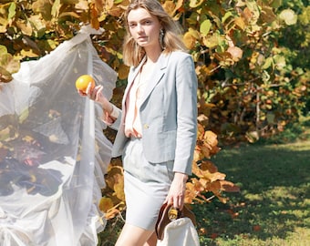 Puede incluir: Una mujer con un blazer y una falda azul claro, sosteniendo una fruta cítrica amarilla en su mano. Está de pie frente a un arbusto con hojas amarillas y una tela blanca transparente.