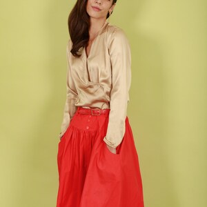 May include: A woman wearing a beige satin blouse with a red skirt and a brown belt. The skirt has a gathered waist and pockets.