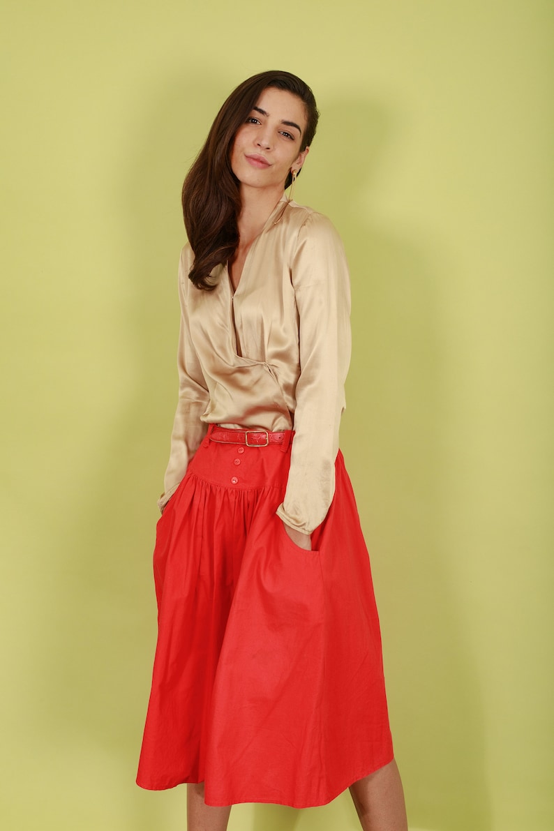 May include: A woman wearing a beige satin blouse with a red skirt and a brown belt. The skirt has a gathered waist and pockets.