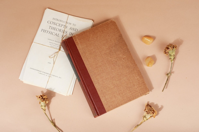 K&ouml;nnte beinhalten: Ein braunes Buch mit rotem R&uuml;cken und ein Stapel wei&szlig;er B&uuml;cher mit dem Titel "Introduction to Concepts and Theories in Physical Solid State" auf einer hellrosa Oberfl&auml;che. Auf der Oberfl&auml;che befinden sich zwei kleine gelbe Steine und zwei getrocknete Blumen.