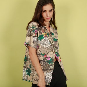 May include: A beige short-sleeved button-up shirt with a colorful floral and map print. The shirt has a relaxed fit and a collar.
