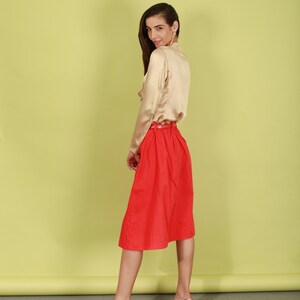May include: A woman wearing a beige satin blouse and a red skirt. She is standing in front of a yellow background and is looking over her shoulder.