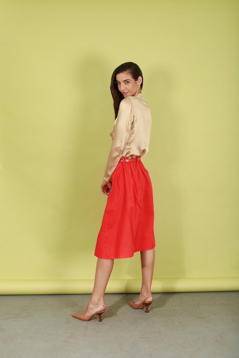 May include: A woman wearing a beige satin blouse and a red skirt. She is standing in front of a yellow background and is looking over her shoulder.