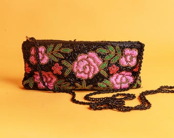 May include: Black beaded clutch with pink and red floral embroidery. The flowers are accented with green leaves. The clutch has a black beaded chain strap and a zipper closure. The background is a solid orange color.