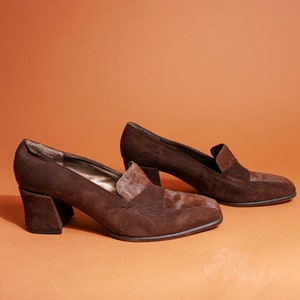 May include: A pair of brown suede heeled shoes with a block heel. The shoes have a pointed toe and a small flap detail on the top.